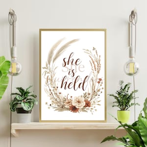 May include: A framed print with the words "she is held" in a brown script font, surrounded by a wreath of beige and brown floral elements. The frame is gold. The print is displayed on a wooden shelf with plants and hanging light fixtures.