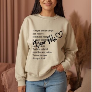 Peut inclure: Un sweat-shirt beige avec le texte "Strength doesn't always look fearless. Sometimes strength is simply showing up... Dear Me." Un cœur noir est à côté des mots "Dear Me."