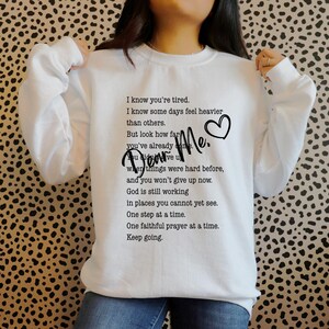 Peut inclure: Sweat-shirt blanc avec le texte "Dear Me." et un message inspirant en noir. Le design comprend un symbole de cœur. Un vêtement décontracté et confortable.