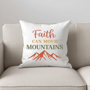 Op de afbeelding: Wit sierkussen met de tekst "Faith CAN MOVE MOUNTAINS" in oranje en groen, met een oranje bergafbeelding. Het kussen ligt op een beige bank.