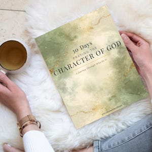 May include: A book titled "30 Days Praying the Character of God" with a watercolor design in shades of green and gold. The book is held on a white fluffy surface next to a gold-rimmed mug.