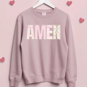 Op de afbeelding: Een mauvekleurige sweatshirt met het woord "AMEN" op de borst. De letters zijn gemaakt van patchworkstof met roze stippen, ruitjes en bloempatronen. De sweatshirt hangt aan een witte hanger tegen een roze achtergrond met hartdecoraties.