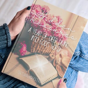 Puede incluir: Un libro titulado "Hidden Treasures: A Year of Deep Diving Into God's Word" con una portada de diseño floral. El libro está abierto en una página con texto. El nombre del autor, "SheSurrendersTruth", está impreso en la parte inferior del libro.