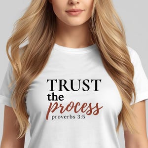 Puede incluir: Camiseta blanca con el texto "TRUST the process proverbs 3:5" en escritura negra y burdeos. La camiseta es de cuello redondo y la lleva una persona con pelo largo y rubio.