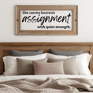 Könnte beinhalten: Ein gerahmtes Holzschild mit dem Text "She carries heaven's assignment with quiet strength." Das Schild ist über einem Bett mit Kissen und einer gemusterten Decke angebracht. Der Rahmen des Schildes ist hellbraun.