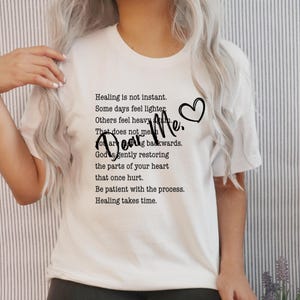 Peut inclure: T-shirt blanc avec le texte "Dear Me." et un cœur. Le t-shirt présente également une citation plus longue sur la guérison et la patience. C'est un vêtement décontracté pour tous les jours.