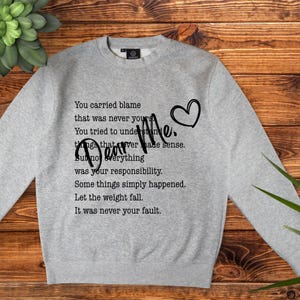 Peut inclure: Un sweat-shirt gris clair avec les mots "Dear Me" et un motif de cœur. Le sweat-shirt a un col rond et présente un poème en texte noir. Le thème du poème est l'auto-compassion et le lâcher prise.