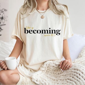 May include: Cream-colored t-shirt with the text "She is becoming" in black and gold lettering, with "psalm 46:5" below. The shirt is worn by a person holding a white mug. The overall theme is inspirational.
