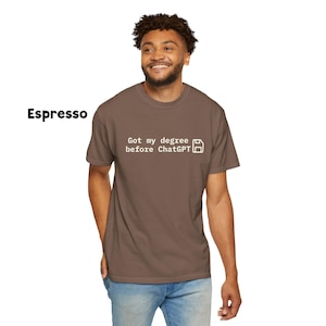 May include: Espresso-colored t-shirt with the text "Got my degree before ChatGPT" in white. The shirt has a classic crew neck and short sleeves. The model is wearing blue jeans.
