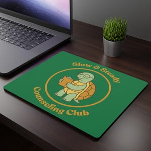 May include: Green mouse mat with gold text reading "Slow & Steady Counseling Club". The design features a cartoon turtle wearing glasses and holding a clipboard. A laptop and small potted plant are visible in the background.