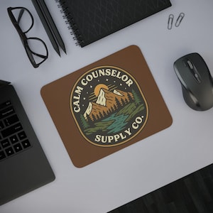 May include: A brown rectangular mousepad with the text "CALM COUNSELOR SUPPLY CO." in a circular design. The design features a mountain range, trees, and a river. A laptop, glasses, pencils, and a mouse are also visible.