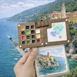 May include: A wooden watercolor palette with 12 paint wells, a water brush, and a small watercolor painting. The palette is open, revealing a selection of vibrant colors and a sample watercolor artwork. A small sketchbook with a watercolor painting is also visible.
