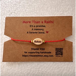 May include: A red string bracelet with a central oval wooden bead engraved with the name Rohan, flanked by two small gold-toned beads. The bracelet is attached to a brown card featuring the text More Than a Rakhi, It’s a promise, A memory, A forever bond, and a QR code.
