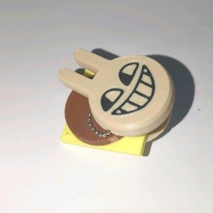 Puede incluir: Un clip de plástico beige con una cara de conejo de dibujos animados con una amplia sonrisa. El mecanismo del clip incluye un disco circular marrón y una base cuadrada amarilla, con una pequeña cadena de bolas de metal plateado en el lateral.