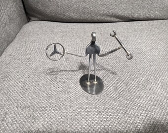 May include: A silver-coloured metal figurine crafted from repurposed cutlery and hardware. The figure stands on an oval base, holding a circular steering wheel emblem in one hand and a small metal wrench in the other, with a fork-like torso.