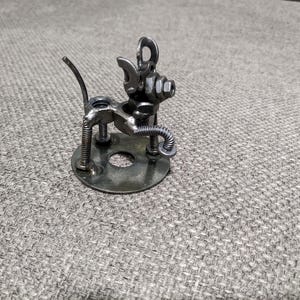 May include: A small metal sculpture of a dog made from nuts, bolts, and other metal pieces. The dog is standing on a round metal base.