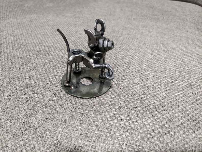 May include: A small metal sculpture of a dog made from nuts, bolts, and other metal pieces. The dog is standing on a round metal base.