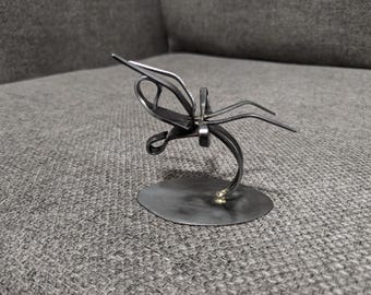 May include: A handcrafted metal sculpture of a dragonfly. The dark grey sculpture is mounted on a circular base and features detailed wings and body. The dragonfly sculpture is a decorative item.