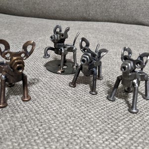 May include: Four metal sculptures of dogs, each made from nuts, bolts, and other metal pieces. The sculptures are standing on a grey fabric surface.