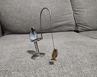 May include: A quirky metal sculpture depicting a fisherman crafted from a fork and other metal components. The fisherman holds a fishing rod with a small fish attached. The sculpture is placed on a grey surface.