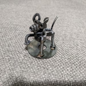 May include: A small metal sculpture of a dog made from recycled metal parts. The dog is standing on a round metal base and has a long tail.
