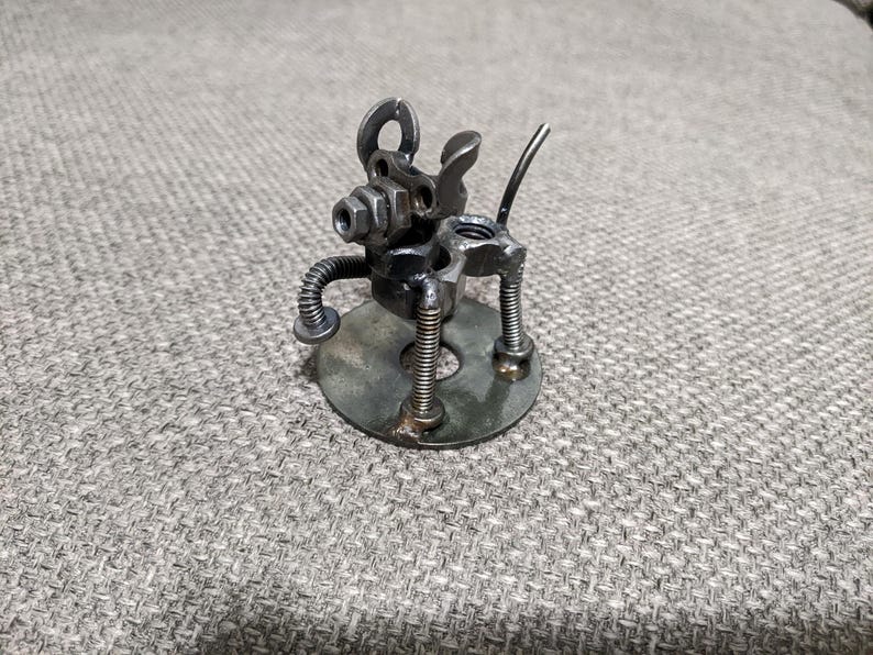 May include: A small metal sculpture of a dog made from recycled metal parts. The dog is standing on a round metal base and has a long tail.