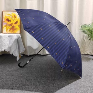 May include: Navy blue umbrella featuring vertical stripes and small gold bow tie motifs. The umbrella has a black handle with a curved hook. The background includes a sunflower painting and a white tablecloth.
