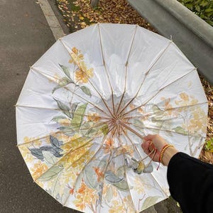 Puede incluir: Parasol blanco abierto con un diseño floral en amarillo, verde y gris. Se ve el marco y el mango de madera. El diseño floral está pintado sobre la tela del paraguas.