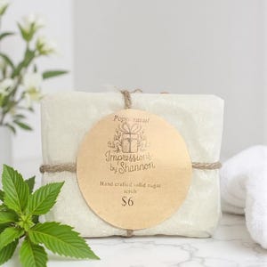 Peut inclure: Un gommage solide au sucre rectangulaire emballé avec une étiquette ronde. L'étiquette indique "Peppermint" et "Impressions by Shannon, Hand crafted solid sugar scrub". Le gommage est attaché avec de la ficelle et a une branche de feuilles vertes.