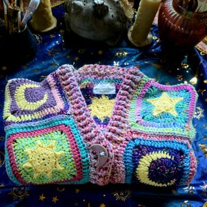 Könnte beinhalten: Eine mehrfarbige Häkeljacke mit quadratischen Feldern, die gelbe Sonnen-, Mond- und Sternmotive zeigen. Das Kleidungsstück hat einen V-Ausschnitt, einen strukturierten Strickrand und einen einzelnen Knopf in Form eines Halbmonds. Etikett Moonlight & Hearth.