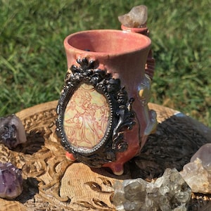 May include: A pink ceramic mug with a black ornate frame featuring a vintage illustration. The mug has a crystal on top and is set on a wooden carved surface with additional crystals. The mug is approximately 4 inches tall.