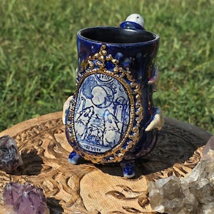 May include: A handmade ceramic mug with a dark blue glaze and gold accents. The mug features a detailed illustration of "The Moon" tarot card within an ornate frame. The mug has small feet and a handle, and is surrounded by crystals.