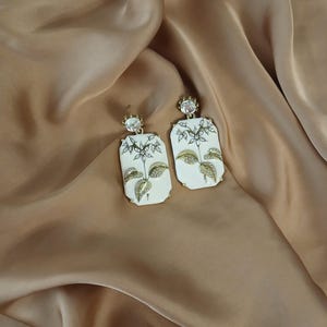 May include: A pair of white and gold earrings with a floral design. The earrings have a rectangular shape with a white background and gold leaf and flower details. The earrings are set against a gold satin background.