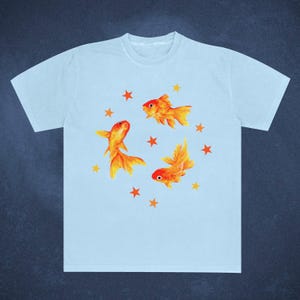 May include: Light blue t-shirt featuring a watercolor design of three orange goldfish and several orange stars. The goldfish have detailed fins and tails. The shirt is a casual, everyday wear item.