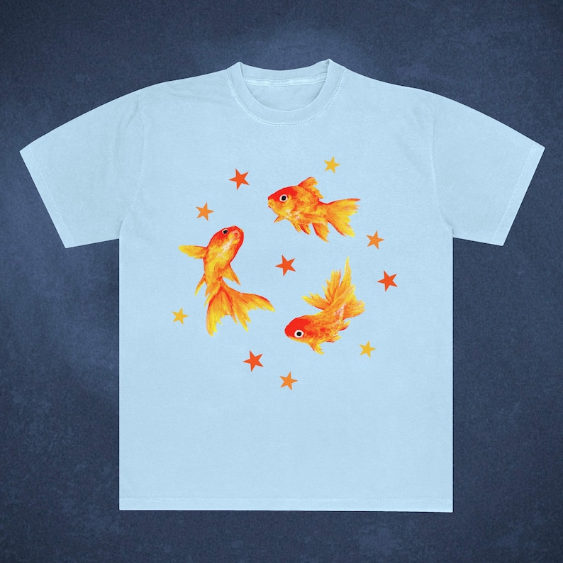 May include: Light blue t-shirt featuring a watercolor design of three orange goldfish and several orange stars. The goldfish have detailed fins and tails. The shirt is a casual, everyday wear item.