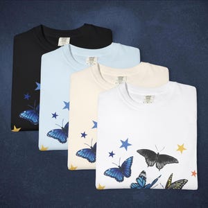 May include: Four folded t-shirts in black, light blue, cream, and white. Each shirt features a butterfly and star design in blue, yellow, and black. The shirts have a crew neck and a tag on the collar.