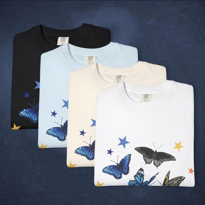 May include: Four folded t-shirts in black, light blue, cream, and white. Each shirt features a butterfly and star design in blue, yellow, and black. The shirts have a crew neck and a tag on the collar.