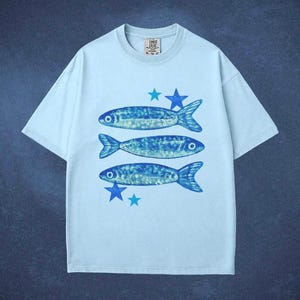 May include: Light blue t-shirt featuring three blue fish and blue star graphics. The shirt has a crew neck and short sleeves. The text "COMET CRUISE" is printed on the collar.