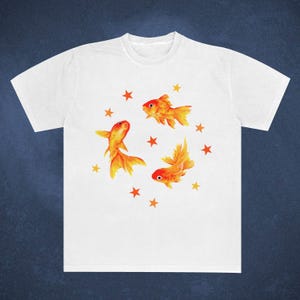 May include: White t-shirt featuring a watercolor design of three orange goldfish and several orange stars. The goldfish have detailed fins and tails, and the stars are scattered around them.