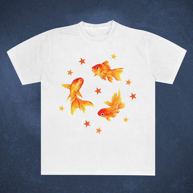 May include: White t-shirt featuring a watercolor design of three orange goldfish and several orange stars. The goldfish have detailed fins and tails, and the stars are scattered around them.
