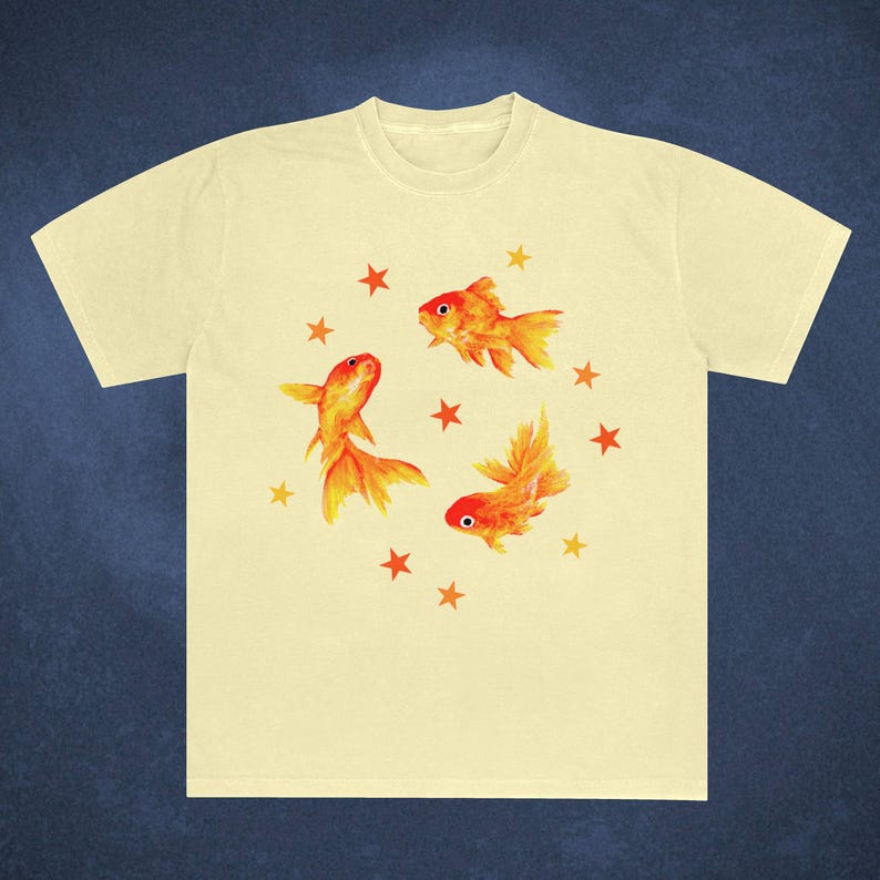May include: A light yellow t-shirt featuring a watercolor design of three orange goldfish and several orange stars. The goldfish have detailed fins and tails, and the stars are evenly spaced around the fish.