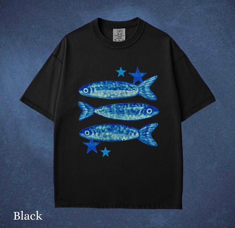 May include: Black t-shirt featuring three blue fish and blue stars. The fish have a textured design. The shirt has a crew neck and short sleeves. The word "Black" is at the bottom of the shirt.