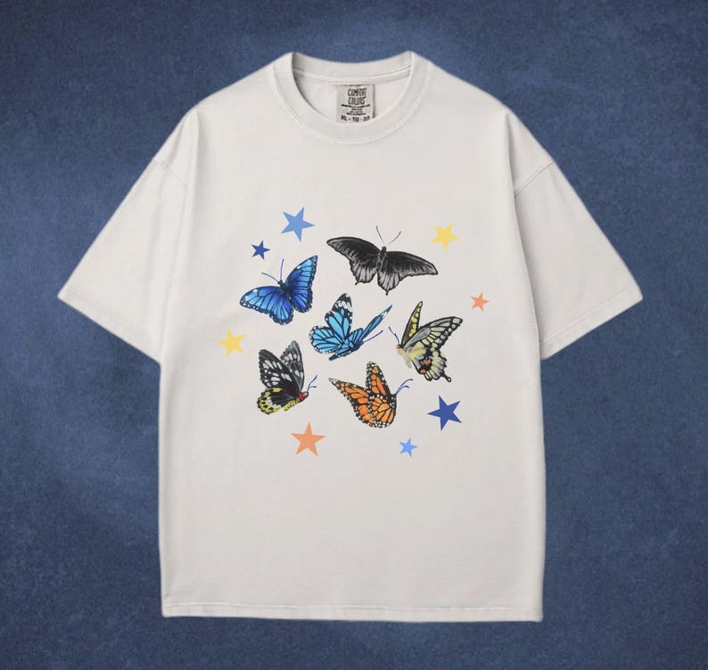 May include: White t-shirt featuring a colorful butterfly and star design. The butterflies are in various colors, including blue, black, and orange, with yellow and blue stars. The shirt has a crew neck and short sleeves.