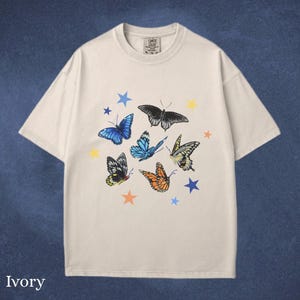 May include: Ivory-colored t-shirt featuring a colorful butterfly and star design. The butterflies are in various colors, including blue, orange, and black. The shirt has a classic crew neck and short sleeves.