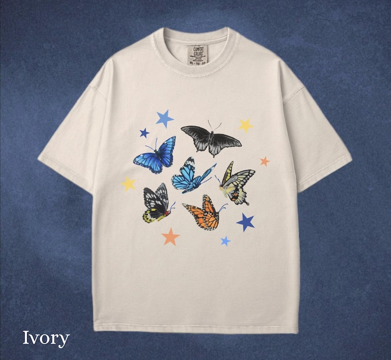 May include: Ivory-colored t-shirt featuring a colorful butterfly and star design. The butterflies are in various colors, including blue, orange, and black. The shirt has a classic crew neck and short sleeves.