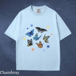 May include: Light blue t-shirt with a butterfly and star graphic. The design features various butterflies in shades of blue, orange, yellow, and black, along with blue and yellow stars. The word "Chambray" is printed at the bottom.