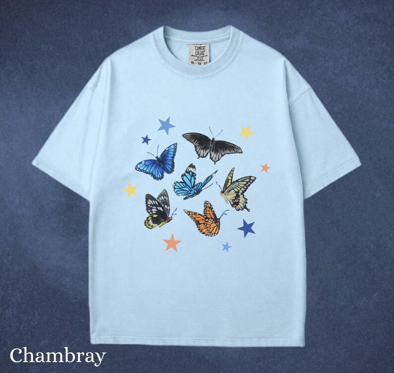 May include: Light blue t-shirt with a butterfly and star graphic. The design features various butterflies in shades of blue, orange, yellow, and black, along with blue and yellow stars. The word "Chambray" is printed at the bottom.