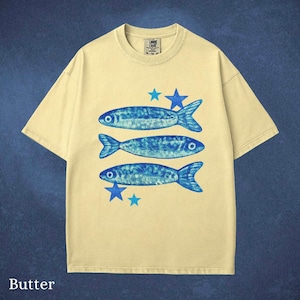 May include: Butter-colored t-shirt featuring three blue fish and blue stars. The fish have a textured design. The shirt has a crew neck and short sleeves. The word "Butter" is printed at the bottom of the shirt.