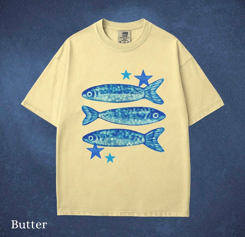 May include: Butter-colored t-shirt featuring three blue fish and blue stars. The fish have a textured design. The shirt has a crew neck and short sleeves. The word "Butter" is printed at the bottom of the shirt.