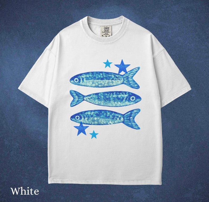 May include: White t-shirt featuring three blue fish and blue star graphics. The shirt has a crew neck and short sleeves. The text "COMET CRUSH" is printed on the upper left of the shirt.